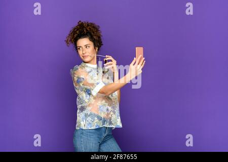 Vue latérale de la brunette positive à poil dur dans la tendance blouse et jean tenant des lunettes de soleil et tenant le selfie sur mobile téléphone en vous tenant à nouveau Banque D'Images