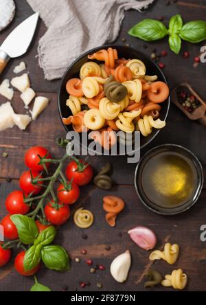 Pâtes de trotole tricolore fraîches et crues dans un bol noir avec tomates cerises et basilic, ail et huile avec parmesan sur fond de bois. Vue de dessus Banque D'Images