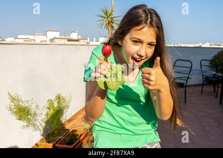 Jolie jeune fille avec son pouce dans une main et un radis fraîchement cueilli dans son jardin urbain dans l'autre. Concept de vie saine Banque D'Images