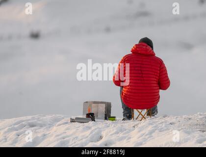 Alpiniste en manteau rouge sur une colline de montagne couverte de neige blanche en faisant une pause pour garder au chaud manger et boire. Vue à flanc de coteau à distance pendant Winte Banque D'Images