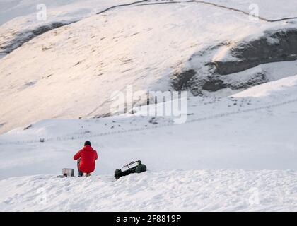 Alpiniste en manteau rouge sur une colline de montagne couverte de neige blanche en faisant une pause pour garder au chaud manger et boire. Vue à flanc de coteau à distance pendant Winte Banque D'Images