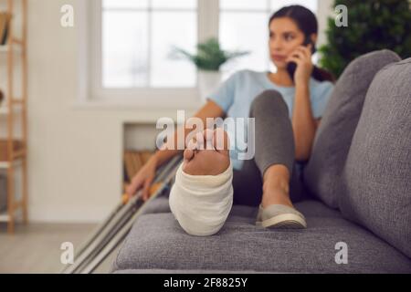 Femme avec une jambe cassée en fonte couchée sur un canapé avec des béquilles à proximité et parler au téléphone Banque D'Images