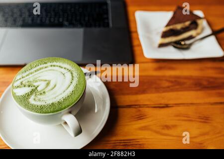 vue sur une tasse de café matcha avec un morceau de gâteau et un ordinateur portable sur une table texturée brune. Bonne mousse sur le café avec un joli motif proéminent. Banque D'Images