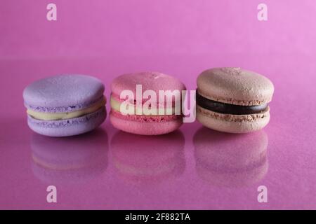 Trois macarons lilas brun chocolat lavande se trouvent dans une rangée sur un fond rose fuchsia-coloré avec réflexion et un lieu pour le texte et l'espace de copie. Banque D'Images