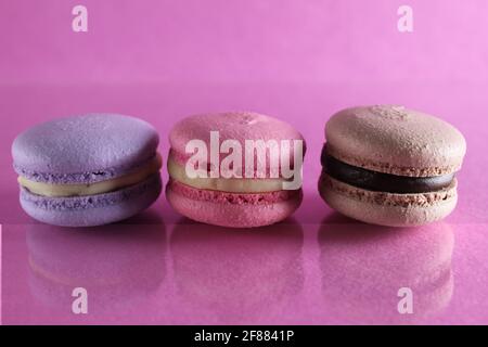 Trois macarons lilas brun chocolat lavande se trouvent dans une rangée sur un fond rose fuchsia-coloré avec réflexion et un lieu pour le texte et l'espace de copie. Banque D'Images