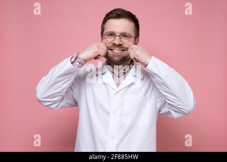 Drôle de médecin dans un manteau blanc fait le sourire artificiel sur le visage avec les mains. Concept de contrainte. Fond rose. Banque D'Images