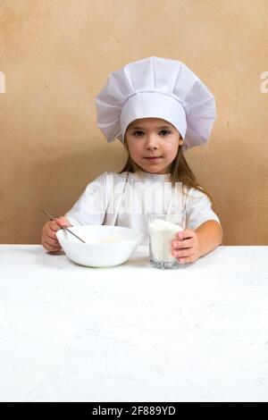 Une petite fille habillée comme un cuisinier pétrit la pâte. Concept de mode de vie enfant de cuisine. Le gamin aime, s'amuse, étudie et joue dans la cuisine Banque D'Images