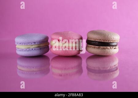Trois macarons lilas brun chocolat lavande se trouvent dans une rangée sur un fond rose fuchsia-coloré avec réflexion et un lieu pour le texte et l'espace de copie. Banque D'Images