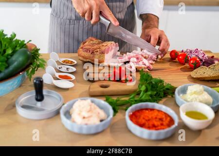 Le chef coupe du bacon dans la cuisine sur une planche à bois. Il y a des légumes, des tranches de pain, des épices et de petits bols de cuisine autour de la surface de travail. Banque D'Images