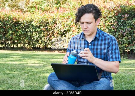 Jeune homme assis dans un parc regardant un ordinateur portable et ouvrant une bouteille bleue réutilisable pour boire de l'eau. Concepts de nomade numérique, travail en extérieur ou étude d'un Banque D'Images