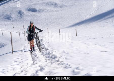 Femme senior en VTT et en train de pousser son e-alpinisme à travers une tache de neige au début du printemps, dans la région d'Allgaeu, en Allemagne Banque D'Images