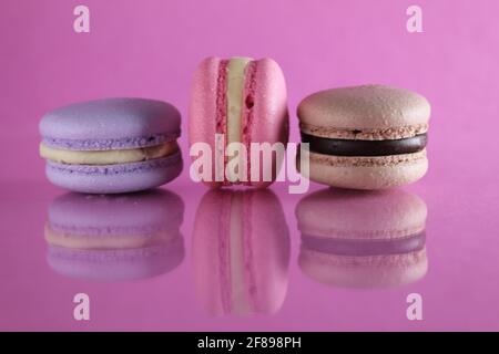 Trois macarons lilas brun chocolat lavande se trouvent dans une rangée sur un fond rose fuchsia-coloré avec réflexion et un lieu pour le texte et l'espace de copie. Banque D'Images