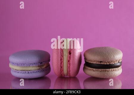 Trois macarons lilas brun chocolat lavande se trouvent dans une rangée sur un fond rose fuchsia-coloré avec réflexion et un lieu pour le texte et l'espace de copie. Banque D'Images
