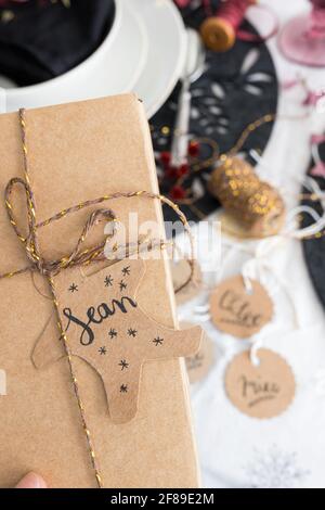 Gros plan sur une boîte cadeau de Noël attachée avec un ruban doré. Le nom Sean figure sur la balise. Plus de tags et une table de dîner élégante est comme flou de dos Banque D'Images