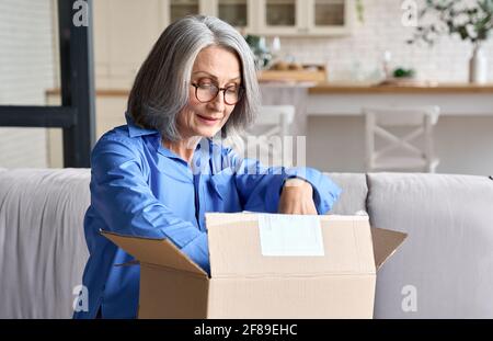 Femme âgée de milieu des années 60 ouvrant la boîte à colis à la maison sur le canapé. Banque D'Images