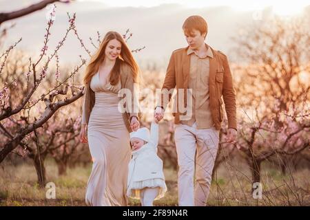 Des promenades en famille dans le jardin rose aux pêches. La jeune mère de père rire, tenir la petite fille par la jeune fille de rouleau à la main par les bras au coucher du soleil. Banque D'Images