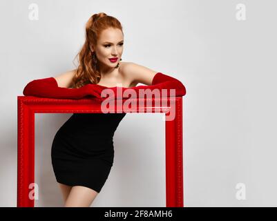 Portrait de la jeune femme artiste en cocktail noir petit à l'épaule robe serrée et gants de longueur coude rouges inclinés sur le cadre rouge Banque D'Images