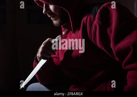 Un homme barbu dangereux dans une cagoule rouge tient un couteau tranchant près de son visage. Un criminel ou un tueur avec des couteaux dans l'obscurité. Banque D'Images