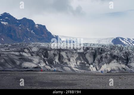 Les personnes en vestes d'extérieur rouges et bleues éclipsent par un glacier lors d'une journée froide et nuageux dans le sud de l'Islande. Banque D'Images