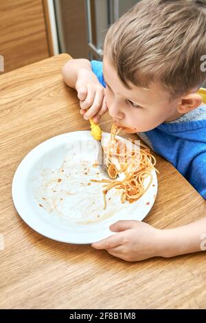 Un petit garçon charmant, avec de grands yeux, mange des pâtes délicieuses et savoureuses avec une fourchette jaune dans la cuisine moderne contre Banque D'Images