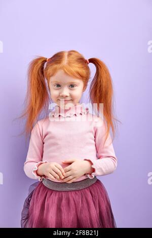 Timide diligent rouge fille avec deux queues poney posant à la caméra, souriant, incroyable fille en jupe isolée sur fond de studio violet Banque D'Images