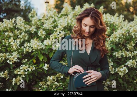 une jeune belle femme adulte dans une robe verte se tient contre un fond de buissons fleuris et tient un petit sac vert dans ses mains. Banque D'Images