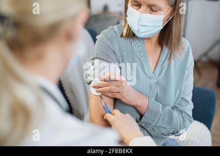 Vue de l'épaule d'une femme médecin méconnaissable. Femme âgée en masque de protection, vaccinée à la maison par son médecin généraliste. Banque D'Images