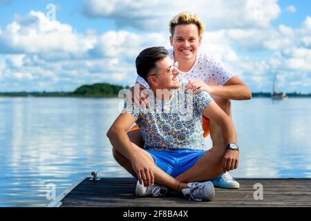 joyeux couple gay souriant assis sur une jetée en bois contre lac et ciel bleu Banque D'Images