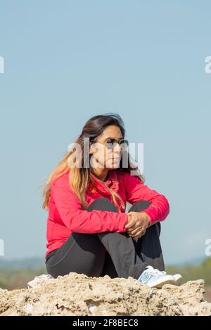 Fille latine, assise sur un rocher de pierre, portant des lunettes de soleil, des bains de soleil et de penser Banque D'Images