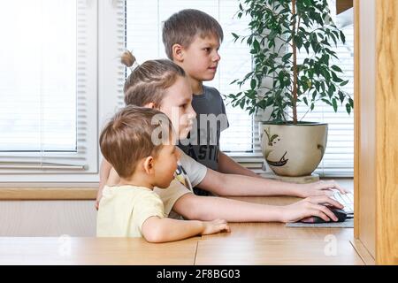 Les écoliers blonds intéressés regardent l'affichage d'ordinateur et les garde d'enfants mains sur la souris et le clavier assis au bureau avec pot usine Banque D'Images