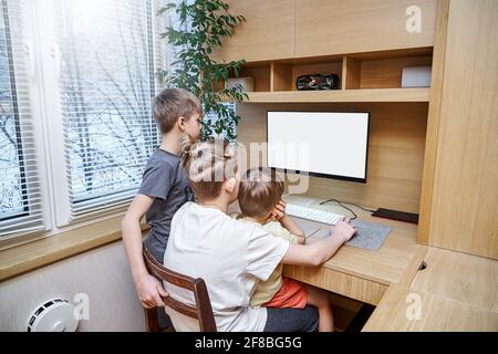 Les jeunes enfants s'assoient sur une chaise au bureau d'ordinateur en bois regarder dans le moniteur blanc près de l'usine de pot vert rester à la maison en quarantaine Banque D'Images