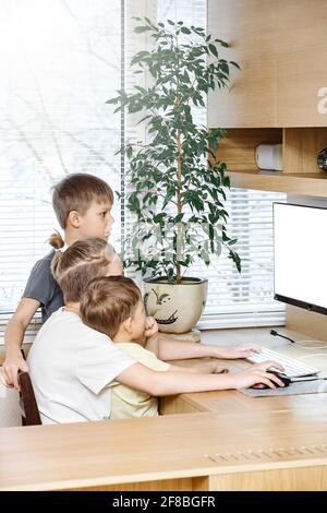 Les jeunes enfants s'assoient sur une chaise au bureau d'ordinateur en bois regarder dans le moniteur blanc près de l'usine de pot vert rester à la maison en quarantaine Banque D'Images