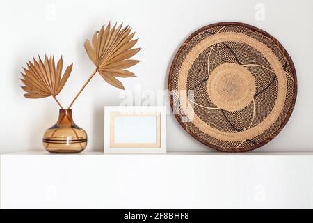 Cadre photo blanc vide à côté d'un vase en verre avec des feuilles de palmier séchées. Banque D'Images