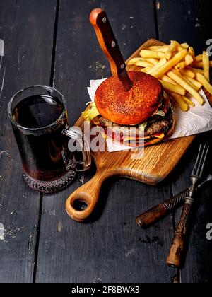 Hamburger de bœuf double appétissant et pommes de terre frites sur une planche à découper en bois, et une tasse de bière foncée. Arrière-plan rustique sombre. Photographie de restauration rapide. Haut v Banque D'Images