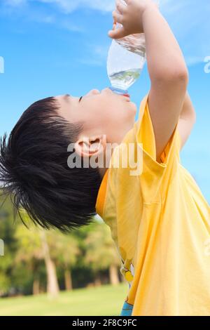 Un petit garçon asiatique boit de l'eau à partir d'une bouteille en plastique avec le soif, après avoir été fatigué d'une poule mouillée dans le parc, sous la lumière du soleil et le ciel bleu Backgrou Banque D'Images