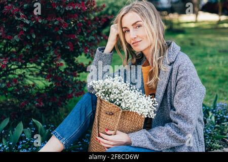 Charmante femme européenne à la mode avec un sac en osier et des fleurs de gitsophila. La fille est assise sur le parapet dans le parc. Portrait de printemps d'un hipster Banque D'Images