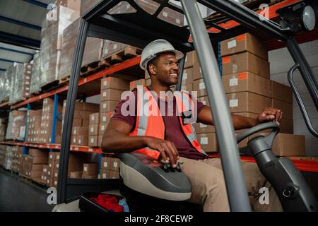 Travailleur ethnique conduisant un chariot élévateur dans un entrepôt où se trouvent les colis de livraison Banque D'Images