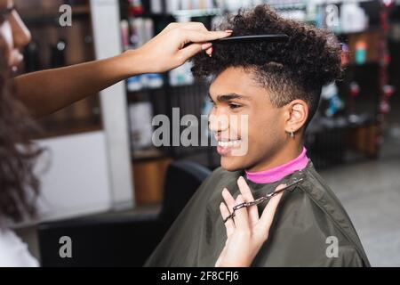 Coiffeur sur le premier plan flou tenant des ciseaux et peignant afro-américain client Banque D'Images