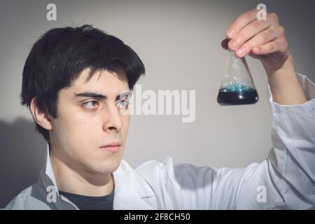 Jeune homme caucasien d'apparence hispanique dans un manteau de laboratoire examinant un échantillon dans une fiole conique, lycée, université, chimie Banque D'Images