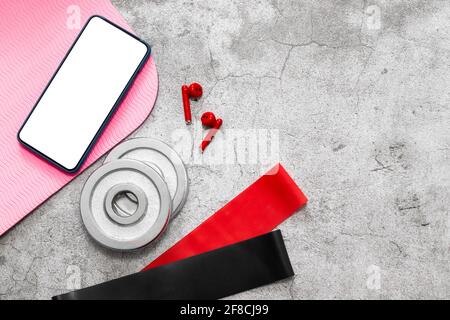 Programme de fitness en ligne. Entraînement en ligne à domicile. Élastiques rouges, tapis de yoga et smartphone sur sol en béton blanc. Banque D'Images
