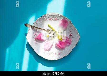 La fleur rose repose sur une assiette vintage, prête à manger. Fourchette rétro argentée. Une affiche avec un design triste inhabituel. Rétroéclairage et jour ensoleillé. Banque D'Images