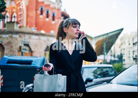 Jeune fille élégante en robe noire avec sacs de shopping est parler au téléphone dans la rue de la ville Banque D'Images