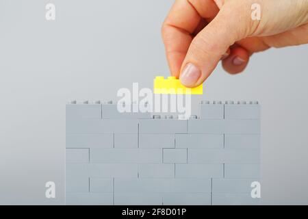Un mur gris fait d'un ensemble de construction de jouet avec la dernière brique jaune en main. Concept Banque D'Images