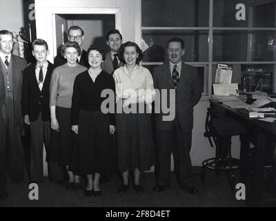 Dans les années 1950, historique, à l'intérieur d'un bureau de l'entreprise, tous ses sourires comme hommes et femmes du personnel de l'administration se tiennent ensemble pour une photo, Angleterre, Royaume-Uni. À cette époque, le lieu de travail au bureau était formel et les employés devaient s'habiller d'une certaine manière. Pour les hommes comme pour les femmes, un costume en laine sur mesure élégant présentait une image professionnelle et aurait été le vêtement approprié dans un environnement de bureau d'entreprise à la fois. Comme on peut le voir, le jeune lad, le bureau junior, porte une veste et une cravate. Banque D'Images