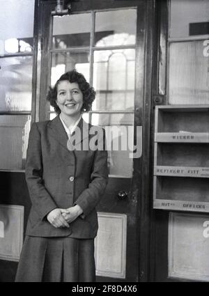 Années 1950, historique, une jeune employée, une secrétaire ou une employée de bureau, debout pour une photo dans son bureau de compagnie, avec un plateau en bois pour les lettres et la correspondance fixé au mur à côté de la porte lambrissée de verre, Angleterre, Royaume-Uni. À cette époque, le lieu de travail au bureau était formel et les employés devaient s'habiller d'une certaine manière. Porter un costume en laine élégant, une combinaison veste/jupe telle qu'elle est, présenterait une image professionnelle et aurait été le vêtement approprié pour une jeune femme dans un environnement de bureau d'entreprise à l'époque, Banque D'Images