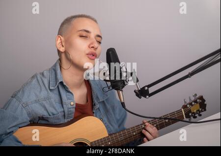 Une jeune femme hipster enregistre une chanson dans un studio d'enregistrement à domicile. La jeune fille joue une guitare acoustique et chante dans un microphone sur fond blanc Banque D'Images