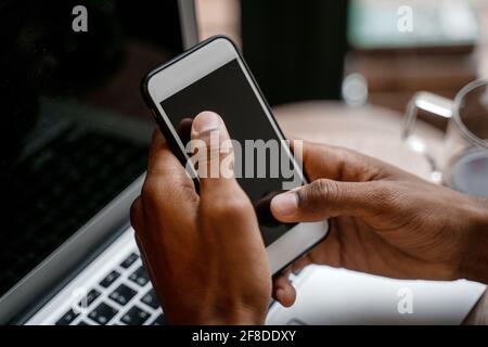 Gros plan du smartphone avec des mains noires sur l'arrière-plan d'un lieu de travail avec un ordinateur portable Banque D'Images