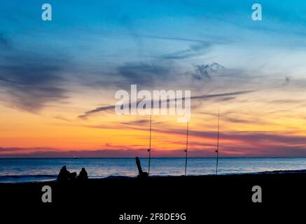 Silhouette de deux personnes assises à la pêche au coucher du soleil sur solitaire plage à l'horizon Banque D'Images