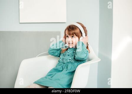 la petite fille de race blanche dans un casque blanc écoute de la musique et danse dans une robe bleu turquoise brillant Banque D'Images