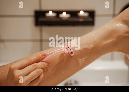 Gros plan jeune femme se raser au rasoir de sa jambe dans le bain confortable rempli de mousse et décoré de bougies. Un traitement et un plaisir sains Banque D'Images